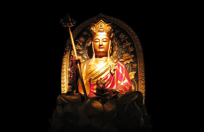 Một vị sư thầy đang ngồi tụng kinh với vẻ mặt trang nghiêm, phía trước là bàn thờ Phật và các vật phẩm thờ cúng, tượng trưng cho nghi lễ cầu siêu và sự thành kính